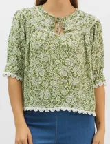 Blusa de gasa estampada con motivos botánicos en color verde, con escote redondo, abertura frontal con lazos de ajuste y mangas cortas. Presenta detalles de encaje en el escote, mangas y ruedo.