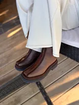 Chaleco tejido de punto en color chocolate, con cuello redondo y diseño sin mangas. Presenta un aplique frontal decorativo compuesto por tres figuras en forma de rombo en tonos tierra, bordeadas con hilo crudo.