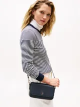 Cartera pequeña de poliuretano color azul marino con acabado liso. Presenta un diseño rectangular con solapa frontal que se cierra magnéticamente y lleva el monograma TH en metal dorado al frente. Incluye correa ajustable y extraíble para llevar al hombro o cruzada.