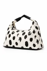 Cartera tipo hobo bag de tela color crema con estampado de manchas negras irregulares. Tiene un asa de hombro de cuero sintético negro y cierre de cremallera.