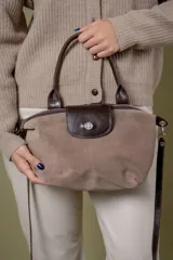 Cartera pequeña de gamuza en tono topo con detalles de cuero marrón oscuro en las asas, solapa frontal y correa. Cuenta con cierre superior y un broche metálico decorativo en la solapa.