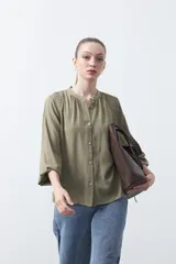 Camisa de color celeste claro con silueta relajada y mangas largas. Presenta un cuello redondo con pequeña abertura frontal y botones, además de un delicado trabajo de tablas en los hombros que aporta textura al diseño.