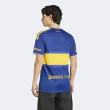 Camiseta de futbol Adidas de la primera equipacion de Boca Juniors, de color azul con la caracteristica franja horizontal amarilla en el pecho. Presenta cuello en V, mangas cortas con las tres tiras de Adidas en los hombros y el logo de la marca y el escudo del club bordados en el frente.