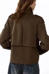 Gabardina corta verde militar de doble botonadura, con solapas amplias, bolsillos laterales diagonales, charreteras en los hombros y mangas raglán amplias.