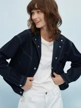 Campera de jean azul claro, estilo trucker, con cuello mao y botones metálicos plateados. Presenta costuras decorativas en el frente y mangas largas con puños doblados.