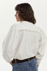 Blusa de algodón liviano color blanco, con detalles de puntilla en el frente, cuello mao con escote en V, cordón colgante y mangas largas abullonadas con puños ajustados.