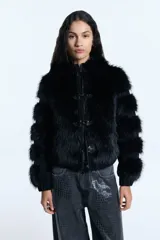 Campera de pelo ecológico negro con recortes en denim negro. Presenta cierre metálico frontal, alamares metálicos decorativos, capucha y bolsillos laterales. Las mangas tienen un diseño con recortes que alternan pelo y denim, y cuenta con elástico en la cintura.