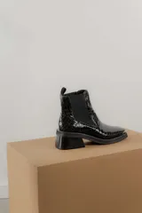 Botas tipo chelsea de cuero color marrón oscuro con acabado brillante. Presentan punta cuadrada con detalle de puntera metálica, elásticos laterales y taco cuadrado bajo.