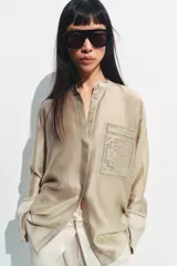 Camisa de cuello mao color beige, confeccionada en tejido satinado con brillo sutil, manga larga con puños anchos y un bolsillo de parche en el pecho con detalles bordados tonales. Cierre frontal con botones ocultos bajo solapa.