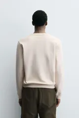 Sudadera de cuello redondo y manga larga en color beige. Presenta un diseño con texto bordado y múltiples parches decorativos en el frente, con terminaciones en rib en puños y cintura.