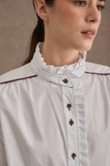 Camisa de popelina blanca con cuello alto tipo victoriano, volados frontales y detalles de costuras y ribetes en color marrón. Presenta mangas largas con puños elásticos terminados en volado y cierre frontal con botones oscuros.
