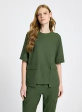 Remera verde militar de tejido de punto con corte recto, cuello redondo y mangas cortas. Presenta bolsillos delanteros alargados con solapas y botones, bajo largo y acabado pespunteado.