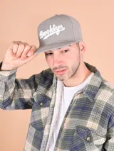 Gorro tipo snapback de color gris claro con visera plana y bordado frontal en blanco con la palabra "Brooklyn" en cursiva.