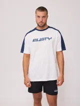 Remera de algodón blanca con mangas raglán en color azul marino y cuello redondo del mismo color. Presenta un estampado frontal con el logo de la marca "Rusty" en tipografía estilizada con efecto 3D en tonos de azul.