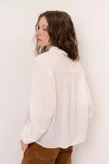 Camisa blanca de manga larga con detalles bordados en el frente, cuello redondo con solapas delicadas, cierre frontal con botones y puños anchos con encaje.