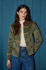 Chaqueta corta de estilo militar confeccionada en terciopelo verde oliva. Presenta cuello mao con detalle en color bordó, cierre frontal con cremallera, botones metálicos decorativos y galones bordados en tono dorado en el frente y puños.