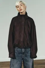 Chaqueta bomber color marrón oscuro con efecto ante, cuello subido y manga larga con puños. Tiene bolsillos delanteros, bajo con pliegues y volumen, forro interior y cierre frontal con cremallera y botones a presión ocultos.