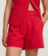 Bermuda sastre de poliéster color rojo, de tiro alto, con cintura asimétrica cruzada, pliegues frontales y bolsillos laterales.