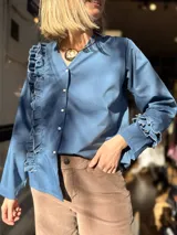 Camisa de popelina color azul, con cuello en V, cierre frontal con botones y diseño de volados laterales y en los puños.