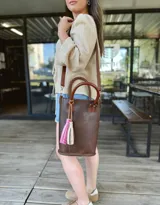 Bolso matera de cuero color marrón con doble asa de mano y correa larga desmontable. Incluye accesorio decorativo de borlas rosas y blancas.