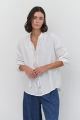 Camisa de color crudo con diseño romántico, cuello redondo con cordón ajustable terminado en borlas, y mangas tres cuartos abullonadas. Presenta detalles de bordados calados verticales en el frente y horizontales en las mangas.