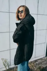 Chaqueta acolchada de color negro con diseño de costuras onduladas, cuello subido y capucha ajustable. Cuenta con cierre frontal de cremallera oculto por una solapa con botones a presión, mangas largas y bolsillos laterales. Tejido con tecnología repelente al agua.