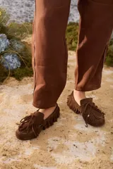 Mocasines de gamuza color marrón, con diseño clásico de mocasín decorado con flecos alrededor y en la capellada, cordón decorativo con avíos dorados y costuras visibles.