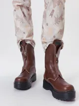 Botas de cuero color marrón de caña alta, con plataforma gruesa de goma y cierre metálico frontal con tirador. Presentan costuras a contratono y detalle de etiqueta de la marca en la lengüeta.
