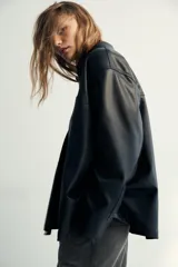 Camisa de efecto piel en color negro, con cuello solapa, manga larga y bolsillos de plastrón en el delantero. Presenta cierre frontal con botones ocultos por tapeta.