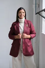 Blazer de corte sastrero en color bordó, con solapas clásicas, bolsillos frontales con solapa y cierre frontal con botones. Presenta una textura sutil en la tela y un broche decorativo en forma de sol en la solapa.
