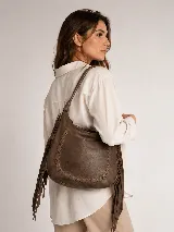 Cartera tipo hobo de cuero vacuno color tostado, con diseño amplio y flexible, decorada con un trenzado artesanal en el frente y largos flecos laterales. Cuenta con correa larga para llevar al hombro.