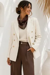 Chaqueta tipo tweed de color marfil o beige claro, con diseño de doble botonadura con botones dorados grabados, solapas clásicas y bolsillos frontales con solapa.