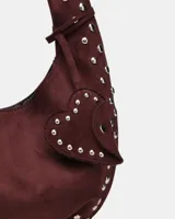 Bolso de hombro tipo hobo color negro, con acabado de gamuza o material similar. Decorado con tachas metálicas redondas plateadas a lo largo del borde inferior y en un dije colgante con forma de corazón.