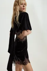 Vestido midi de terciopelo negro con cuello redondo y pañuelo integrado en el mismo tejido. Presenta mangas cortas, aberturas laterales con detalle de encaje a tono y bajo asimétrico con terminación en encaje.