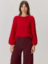 Buzo de lana color beige con mangas abullonadas de tejido calado con diseño de rombos. Presenta un cuello redondo y un detalle de flor negra de tela con cintas colgantes en el escote.