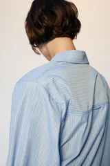 Camisa de manga larga con cuello solapa y hombreras. Presenta un diseño de rayas verticales en tonos azul y blanco, con cierre frontal de botones y puños anchos.