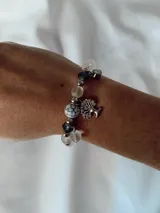 Pulsera elástica compuesta por cuentas de cristal transparente, cuentas oscuras facetadas y una cuenta central con diseño de mármol blanco y negro. Incluye un dije colgante en forma de árbol de la vida plateado y un pequeño dije de cristal.