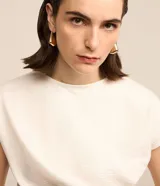 Blusa de manga corta color blanco hueso, con cuello redondo y un detalle de cruce o drapeado asimétrico en la parte delantera a la altura de la cintura. La tela presenta una textura sutil.
