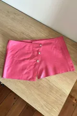 Short pollera rosa de cuero sintético con cierre cruzado y tres botones plateados.