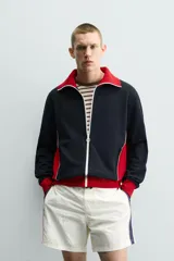 Chaqueta deportiva de corte regular, confeccionada en tejido piqué con interior forrado en red. Presenta un diseño color block en negro y rojo con detalles en blanco, cuello subido, cierre frontal con cremallera y bolsillos laterales con cierre.