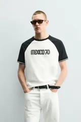 Camiseta de corte regular con diseño color block, cuerpo en color blanco y mangas ranglán en color negro. Presenta cuello redondo, detalles de rib a contraste en los puños y estampado frontal con la leyenda Mexico 70.