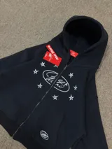 Buzo con cierre frontal, color negro, con estampado gráfico de estrellas y logo en el pecho. Presenta capucha, bolsillos tipo canguro y puños elásticos.