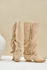 Bota texana alta de gamuza color beige, con taco cuadrado, flecos largos colgantes en el lateral y tiras con hebilla metálica alrededor del tobillo.