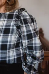 Blusa de manga larga con estampado a cuadros en tonos marrones y grises. Presenta un diseño con volados en los hombros, mangas abullonadas y puños elásticos con terminación de volado.