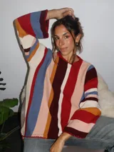 Sweater tejido a mano con terminaciones en crochet, de silueta oversize y mangas acampanadas. Presenta un diseño de rayas verticales multicolores en tonos tierra, rojo, azul y crema.