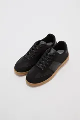 Zapatillas urbanas de color negro con suela de goma marrón, cordones negros con detalles blancos y paneles laterales.