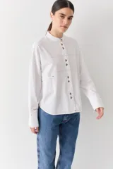 Camisa de corte recto y holgado, color blanco, con cuello mao y cierre frontal de botones. Presenta costuras a la vista en color contrastante, bolsillo lateral tipo parche y puños anchos con detalle bordado.