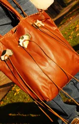 Cartera de cuero color camel, de silueta amplia y blanda, con manijas largas y terminación artesanal en los bordes. Cuenta con apliques decorativos frontales de alpaca y nácar, tientos largos colgantes y bolsillo interno.