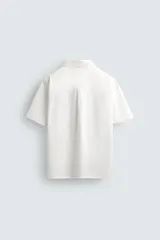 Camisa de manga corta con cuello bowling, confeccionada en popelín de algodón. Presenta un corte relajado, cierre frontal con botones y aberturas laterales en el bajo.