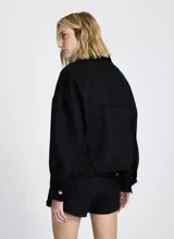 Chaqueta corta negra de tela tejida con cuello alto, mangas largas con puños abotonados, dos bolsillos delanteros con solapa y cierre frontal con botones.